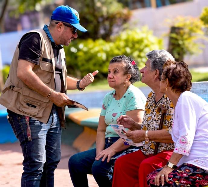 Trabajador de Ibagué Limpia conversa con tres mujeres adultas mayores sentadas en un parque, mientras les entrega volantes informativos.